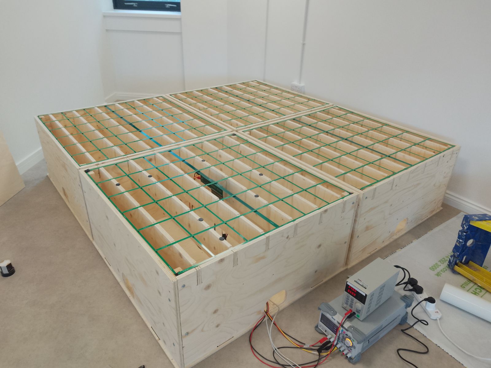 The four plywood quadrants of the 2 m by 2 m antenna floor, top surface removed to show the grid of green coil windings, with a bench power supply and oscilloscope beside it